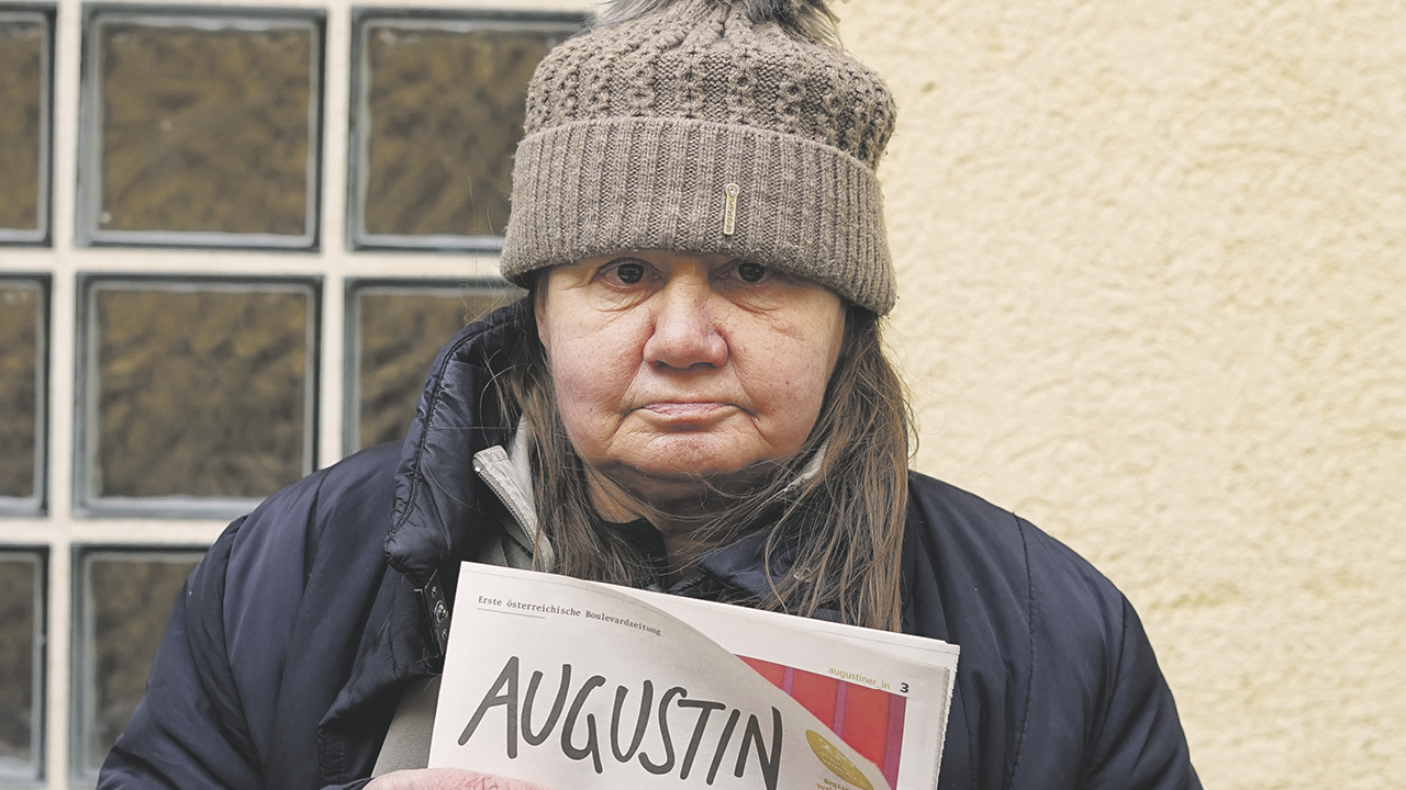 Frau mit tief ins Gesicht gezogener grauer Mütze und Straßenzeitung "Augustin" in der Hand