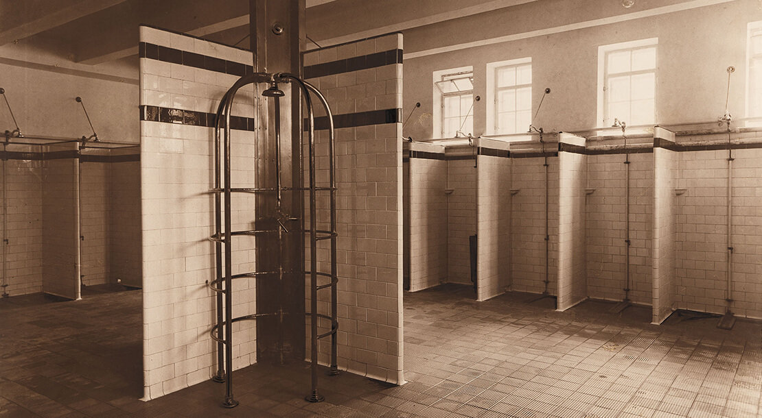 Schwarz/weiß Foto, Innenansicht einer öffentlichen Badeanstalt um 1900, Duschbereich mit gefliesten, nach vorne offenen Einzelduschen. An der längsseitigen Wand Oberlichten
