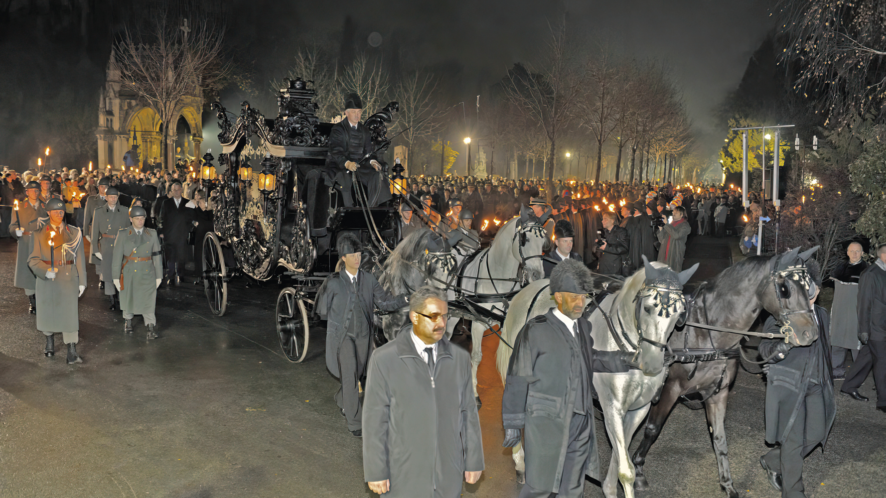 Trauerzug mit schwarzer, prunkvoller Kutsche, die von vier Pferden gezogen wird. Viele Menschen säumen den Zug mit Kerzen in der Hand